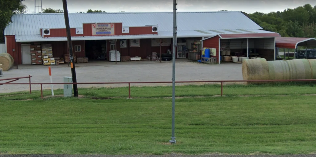 Red metal farm building with metal roof and covered storage areas, hay bales beside metal fence and grass field in foreground